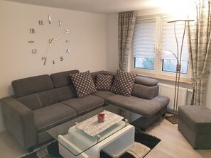 Een moderne woonkamer met een grijze bank, gestreepte kussens, een glazen salontafel en een wandklok.
