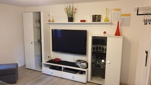 Ett vardagsrum med en platt-TV, en vit TV-bänk, ett glasskåp med vinglas och ett litet bord med en meny.