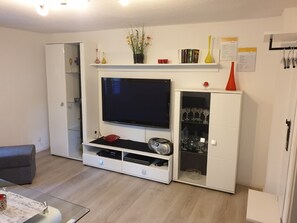 Een woonkamer met een flatscreen televisie, een witte tv-meubel, een glazen kast met wijnglazen en een kleine tafel met een menukaart.