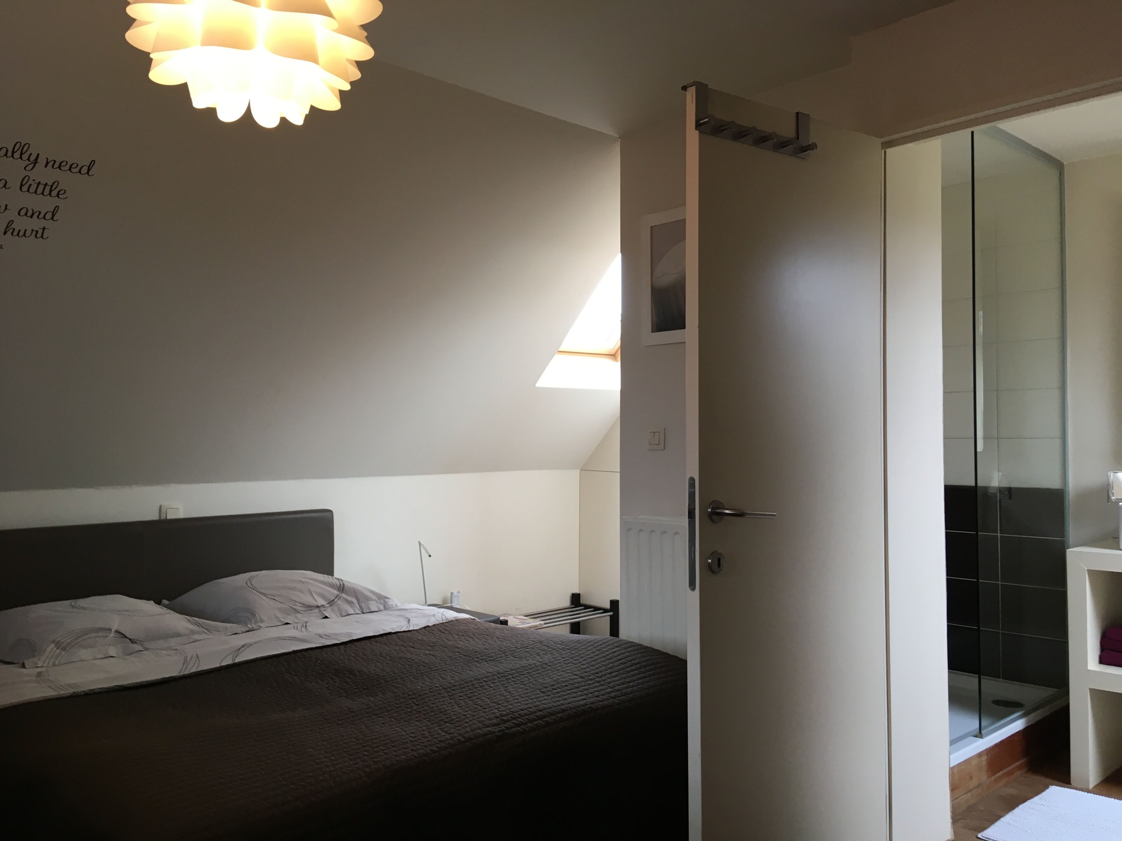 A bedroom with a bed, a bedside table, a nightstand, a wall-mounted light fixture, and a door leading to a bathroom with a shower and a glass-enclosed toilet.