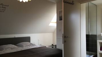 A bedroom with a bed, a bedside table, a nightstand, a wall-mounted light fixture, and a door leading to a bathroom with a shower and a glass-enclosed toilet.