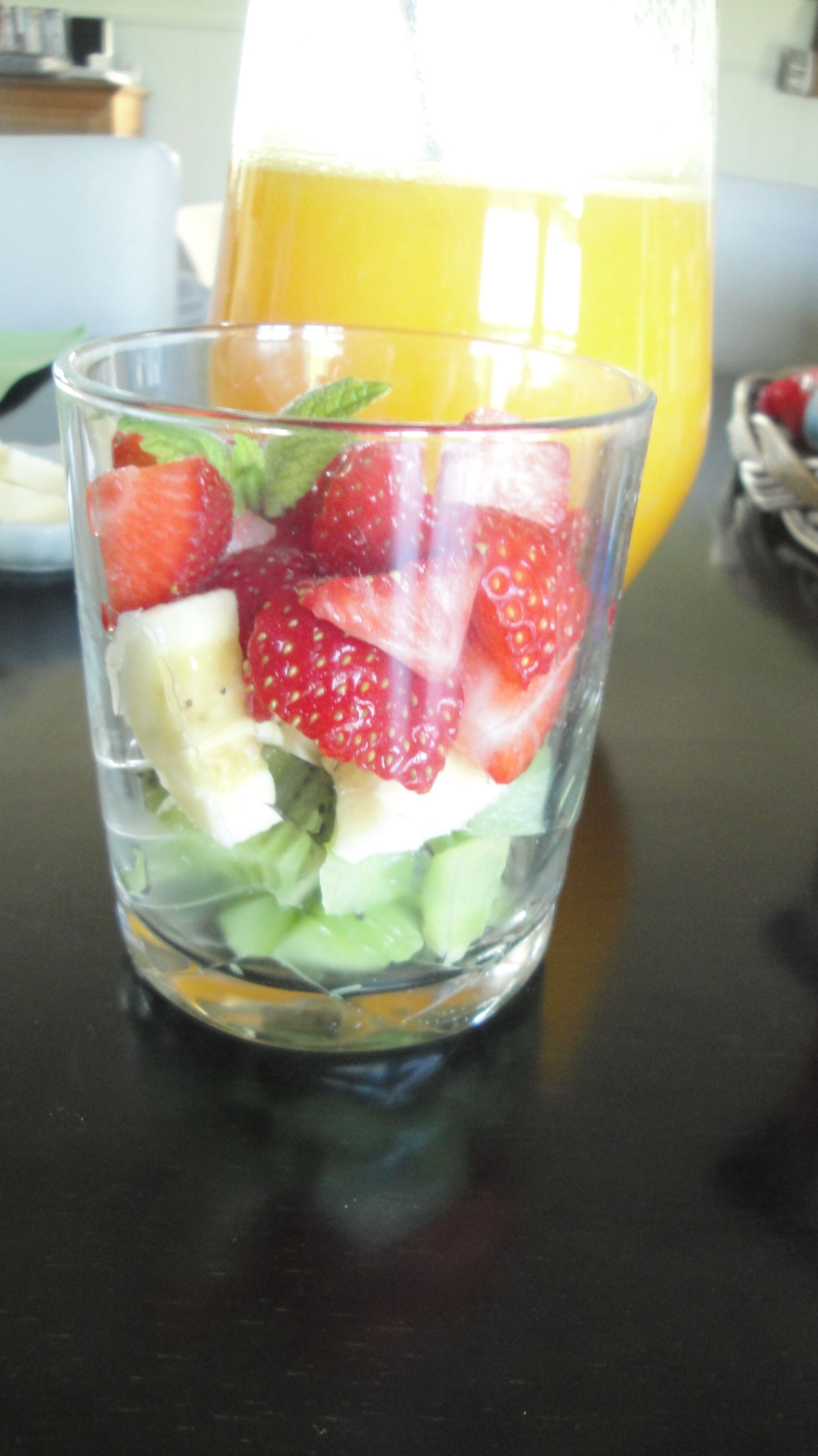 A clear glass containing a fruit salad with strawberries, bananas, and mint leaves.