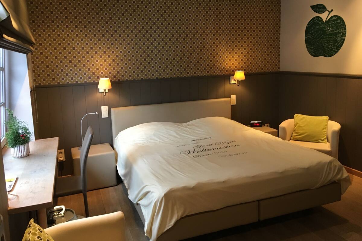 Habitación de hotel con una cama grande, dos lámparas de noche, una mesita con una planta y una pared con un diseño de frutas.