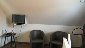 A room with a television mounted on a white wall, two grey chairs, a small wooden table with a remote control, and a radiator on the floor.