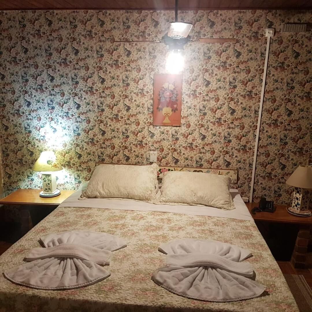A hotel room with floral wallpaper, a bed with two pillows, a nightstand with a lamp, and a picture on the wall.