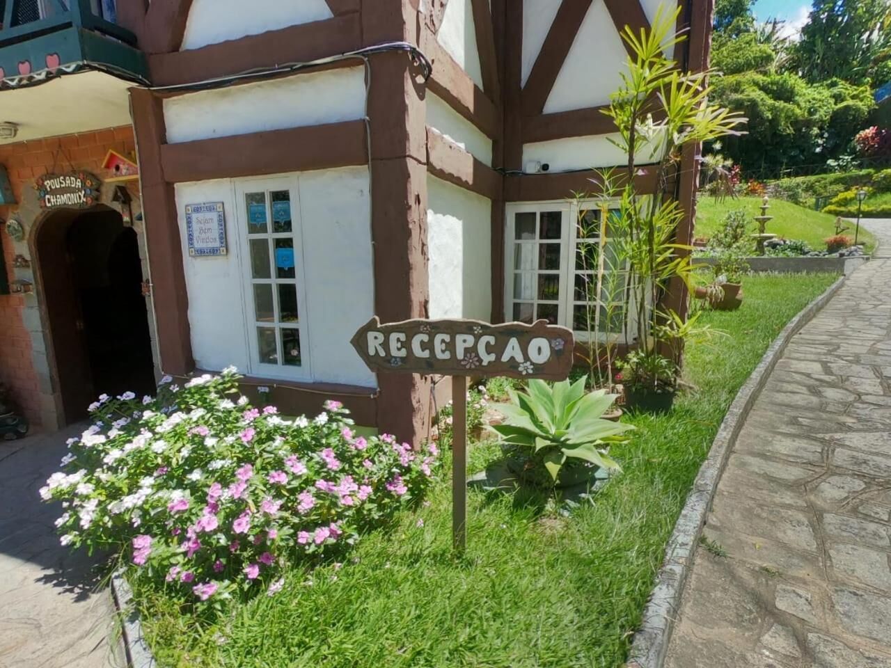 Un edificio con un letrero que dice 'Recepção', un camino de piedra y un jardín con flores rosas.