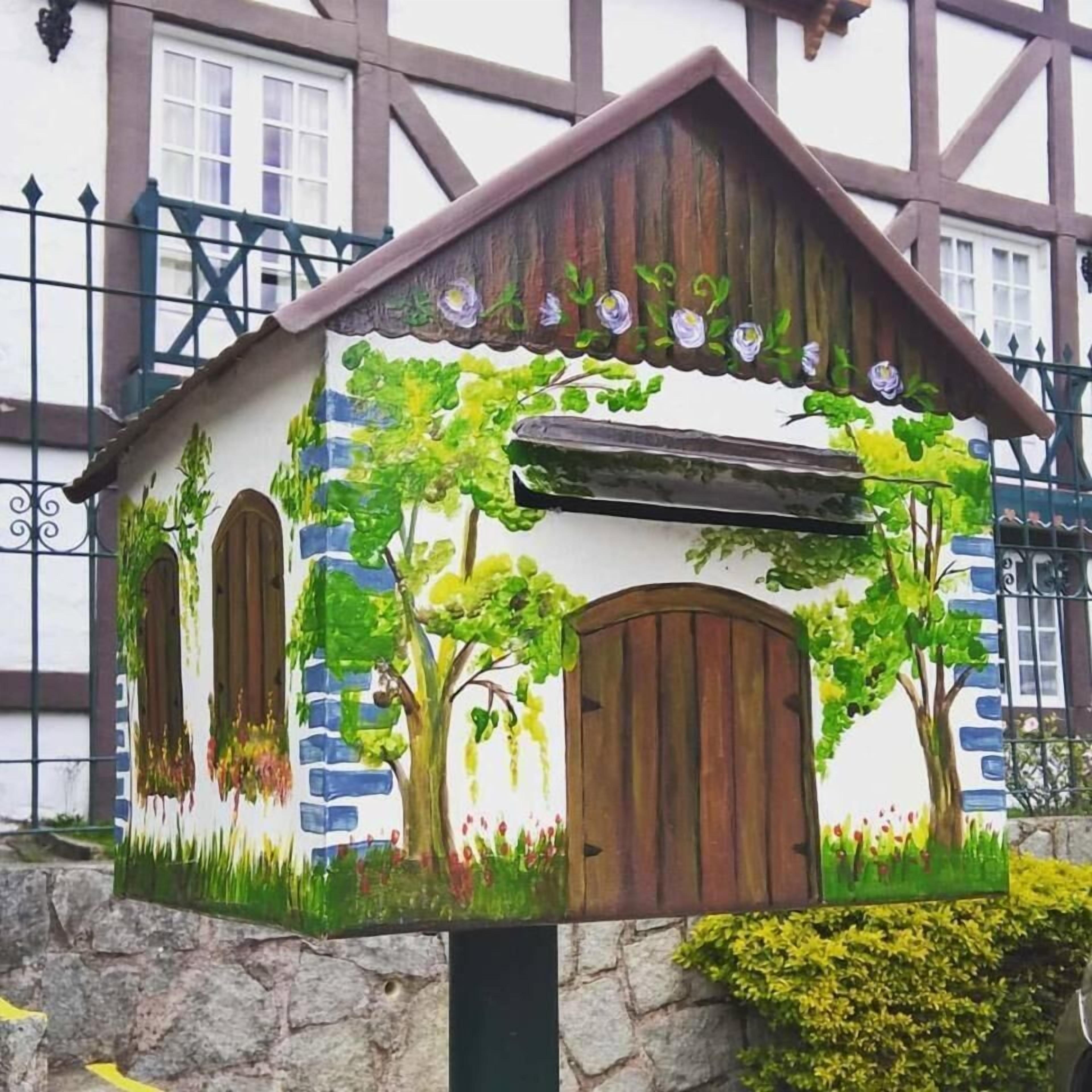 Uma caixa de correio em formato de casa, com exterior pintado, teto marrom, vegetação verde e base de pedra.
