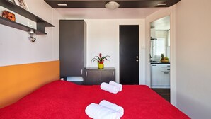 A hotel room with a red carpet, a bed with white towels, a small table with a potted plant, and a kitchenette visible through an open door. - Superior Mobile Homes Kastanija - Campsite (Novigrad)