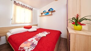 A bedroom with a red bedspread, white pillows, a red heart-shaped pillow, a patterned throw, a shelf with decorative fish, and a potted plant. - Camping Adria Mobile Homes Park Umag (Brtonigla)