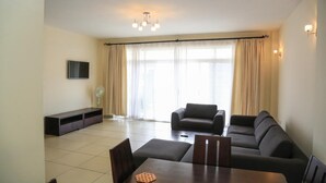 Een woonkamer met een bank, een eettafel met stoelen, een flatscreen televisie aan de muur en een groot raam met gordijnen.