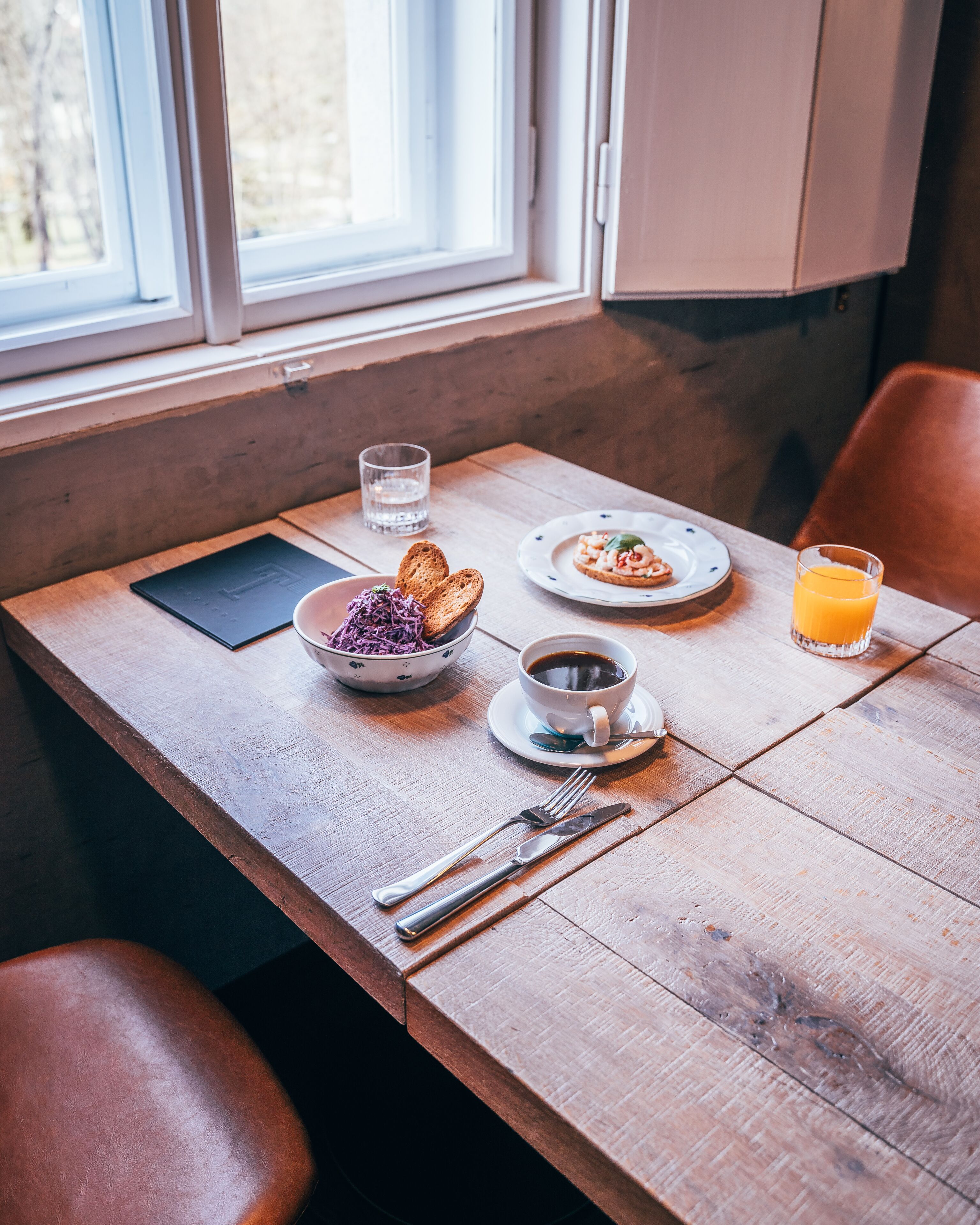 Une table en bois sur laquelle sont disposés des aliments pour le petit-déjeuner, notamment du pain, un saladier rempli de salade, une tasse de café et un verre de jus d’orange.