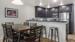 A modern kitchen with dark cabinetry, a stainless steel refrigerator, a microwave, and a dining area with a wooden table and chairs. - Moab Adventure Condo Near Arches | Pool & Hot Tub (Moab)