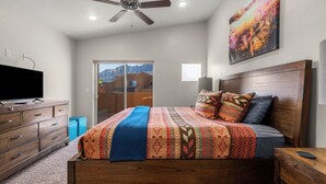 A bedroom with a bed, wooden headboard, a ceiling fan, a TV, a painting, and a window. - Moab Adventure Condo Near Arches | Pool & Hot Tub (Moab)