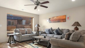 A living room with two sofas, a coffee table, and a rug. There is a ceiling fan, a painting on the wall, and a window with a view of a house. - Redrock Retreat by National Parks | Pool & HotTub (Moab)