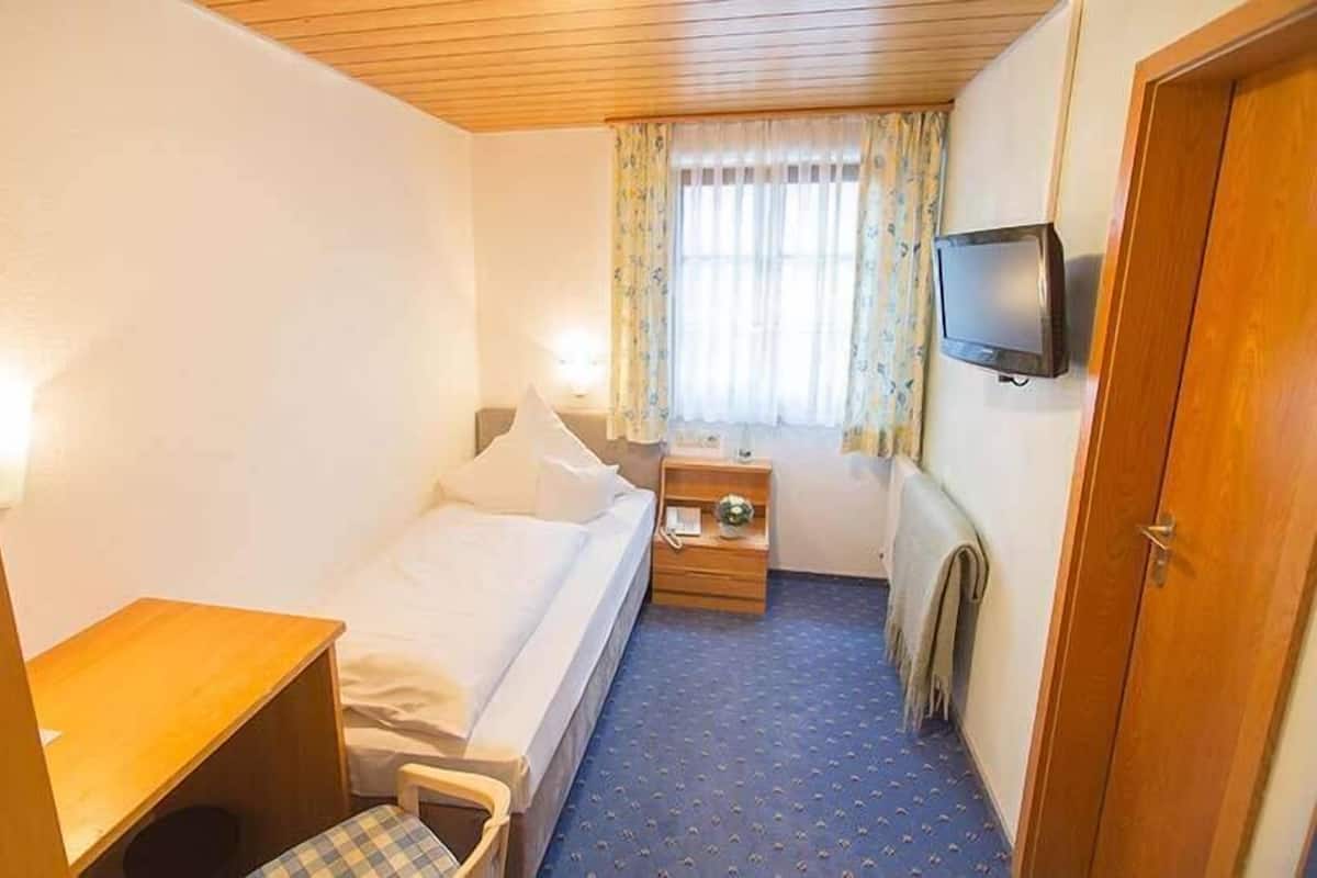 Een kleine hotelkamer met een enkel bed, een bureau, een stoel, een televisie en een raam met gordijnen.