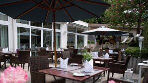 An outdoor dining area with wicker furniture and umbrellas, surrounded by glass windows and potted plants.