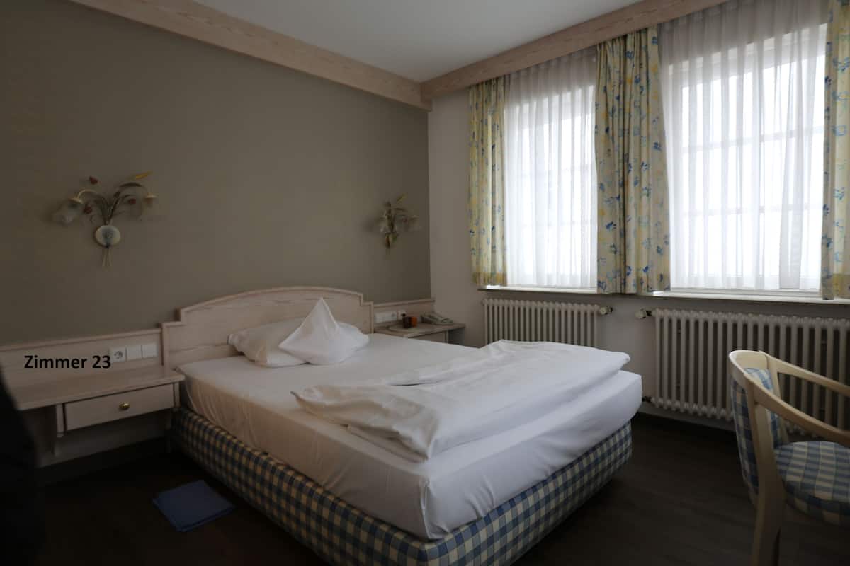 Een hotelkamer met een groot bed, nachtkastje, stoel en een raam met gordijnen.