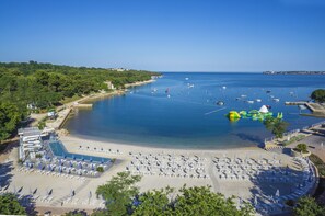 Un complejo turístico frente al mar con una gran piscina, zona de playa con tumbonas y un puerto deportivo con barcos.