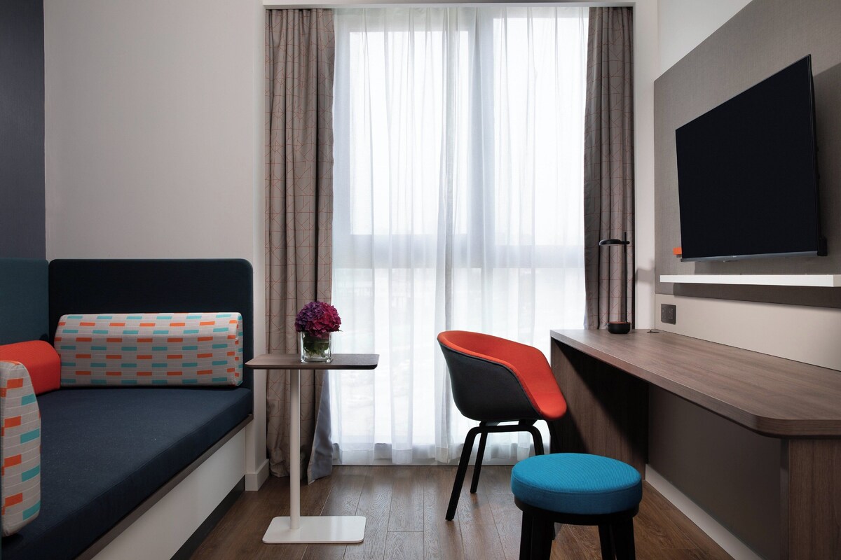 Una habitación de hotel moderna con un sofá, una mesita con un florero, una silla, un taburete y un televisor montado en la pared.