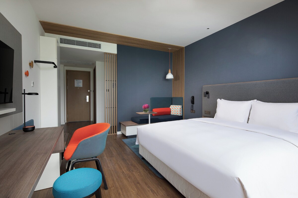 Habitación de hotel con una cama grande, un escritorio con silla, una mesita con lámpara y una puerta que da a otra habitación.