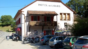 A two-story building with the sign "HOTEL DE LOURS" on the facade. There are several parked cars and motorcycles in front.
