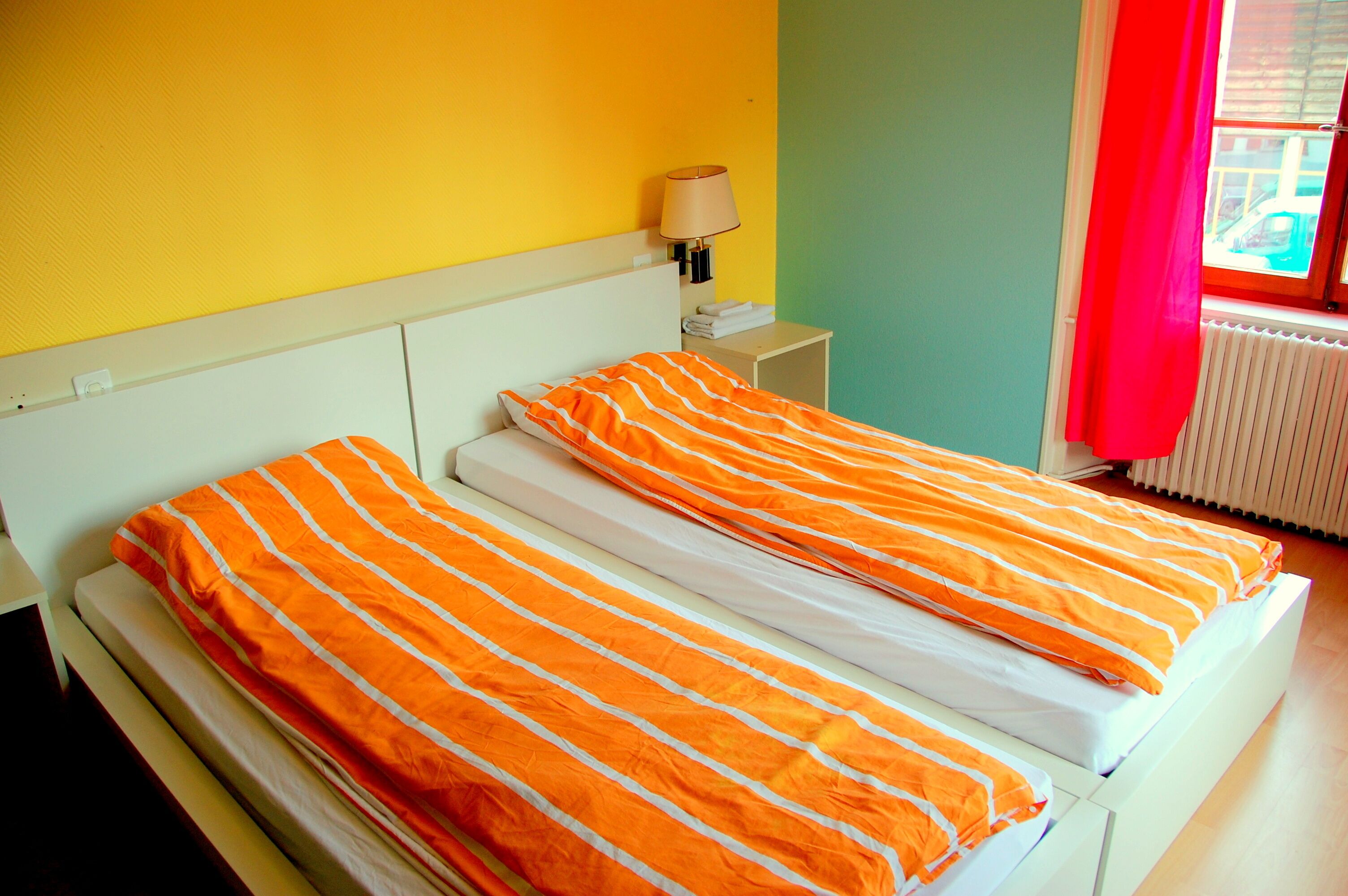 A room with two beds, a nightstand with a lamp, and a window with a red curtain.