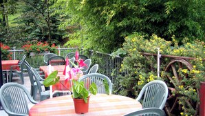 An outdoor patio with round tables and chairs, surrounded by lush greenery and flowering plants.