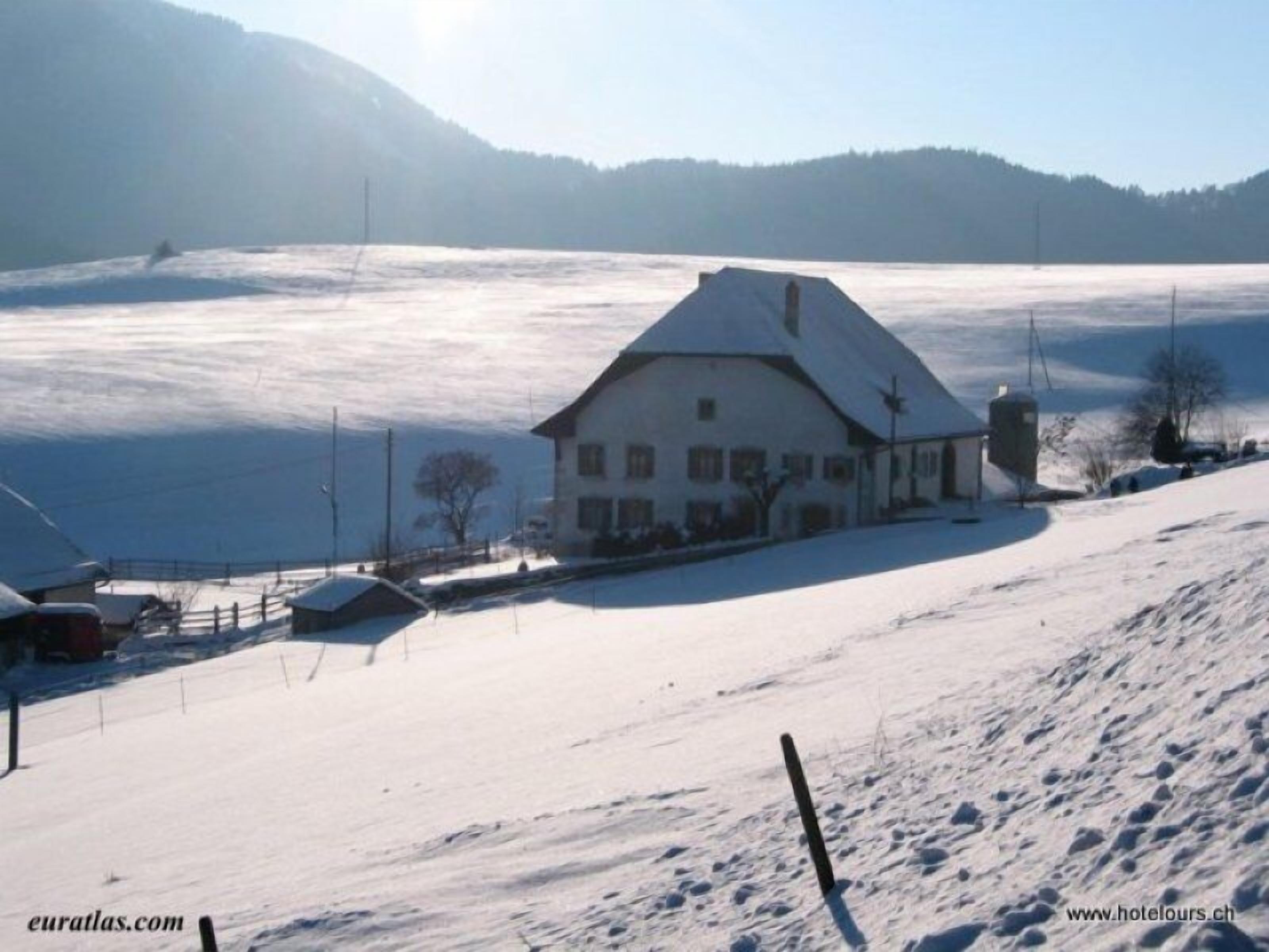 A snow-covered landscape with a white building that appears to be a hotel, surrounded by a snowy field and a mountain in the background.