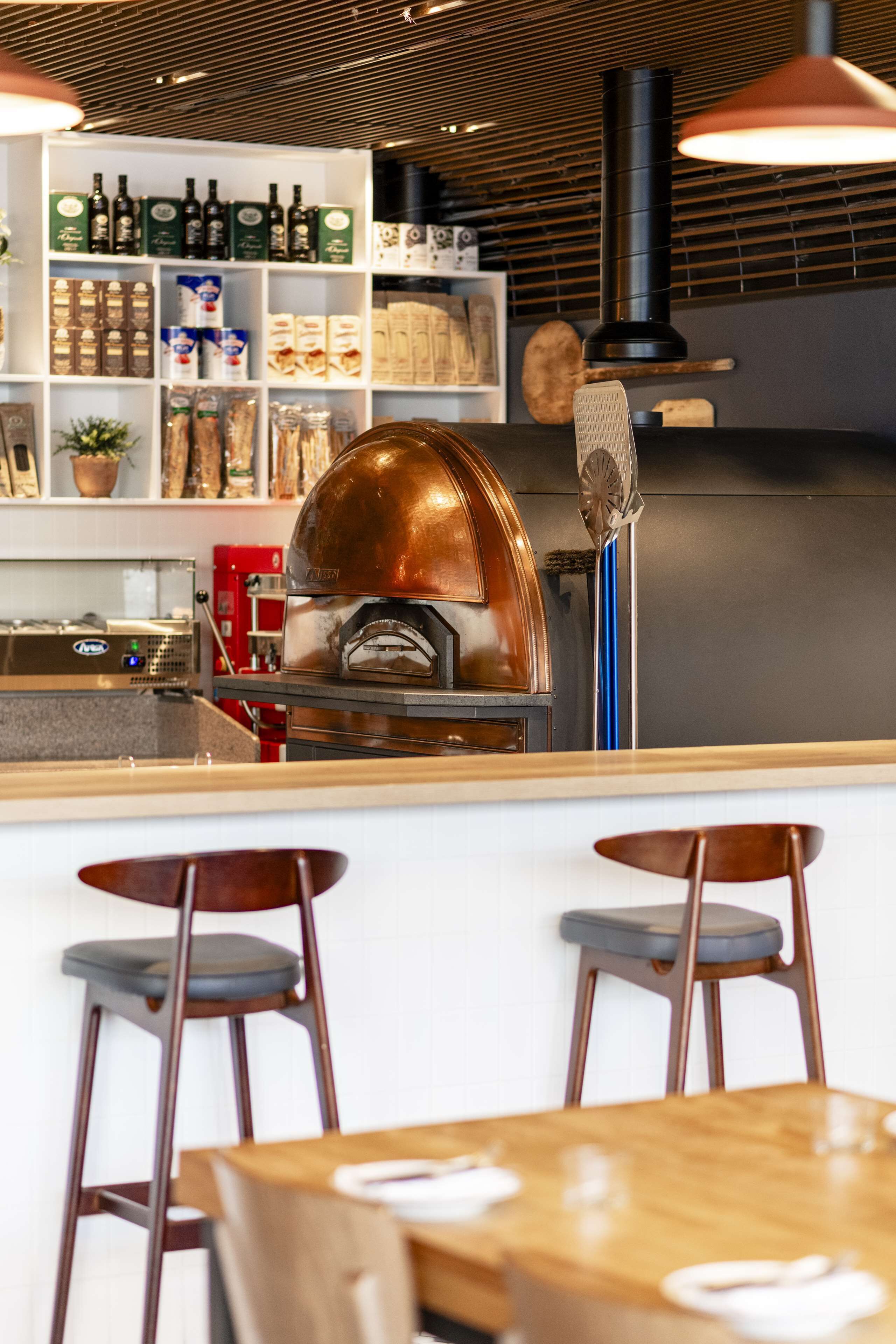 A restaurant interior with a wood and metal bar, two stools, and a pizza oven.