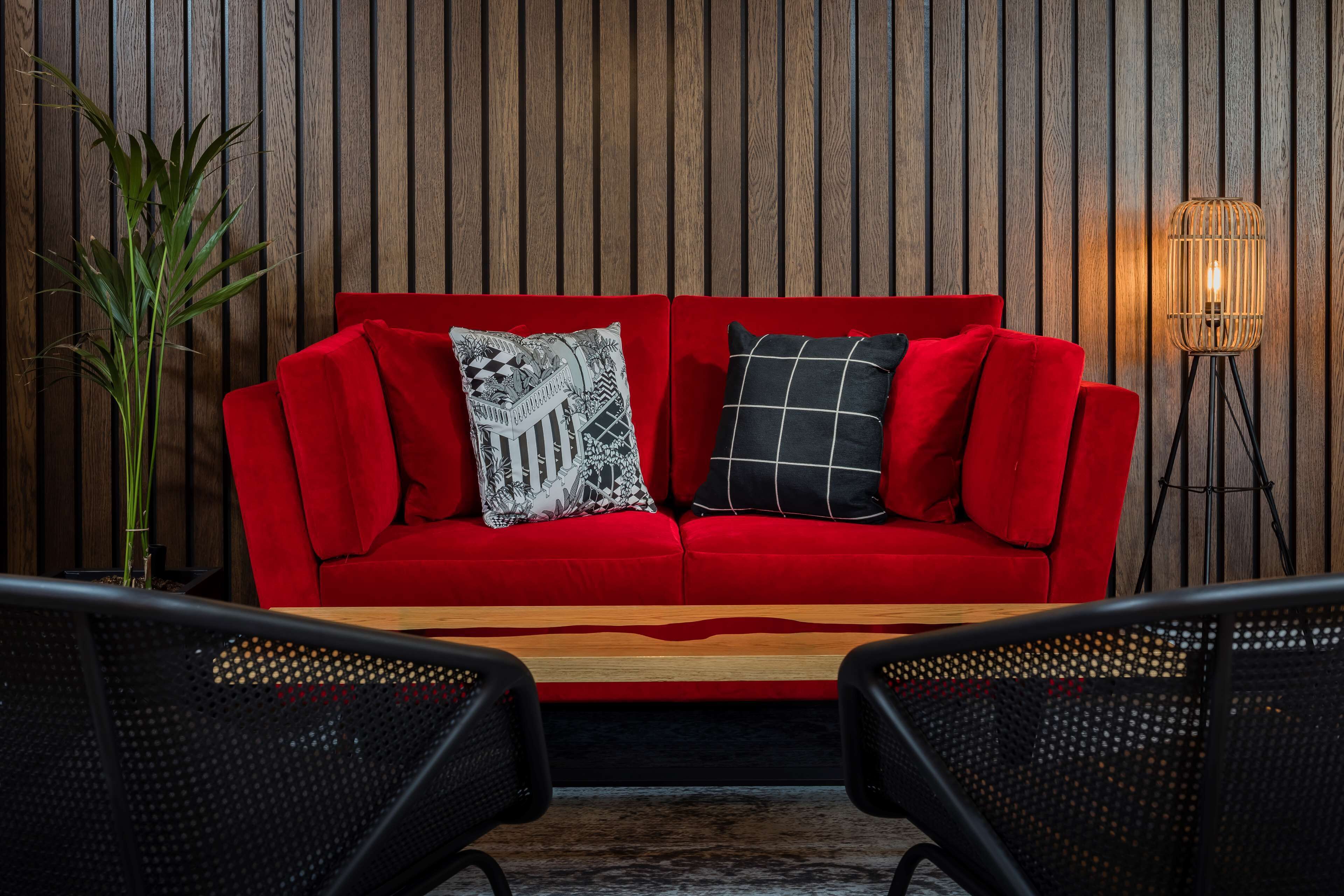 A red sofa with patterned cushions against a wooden wall, accompanied by a floor lamp and two black chairs.