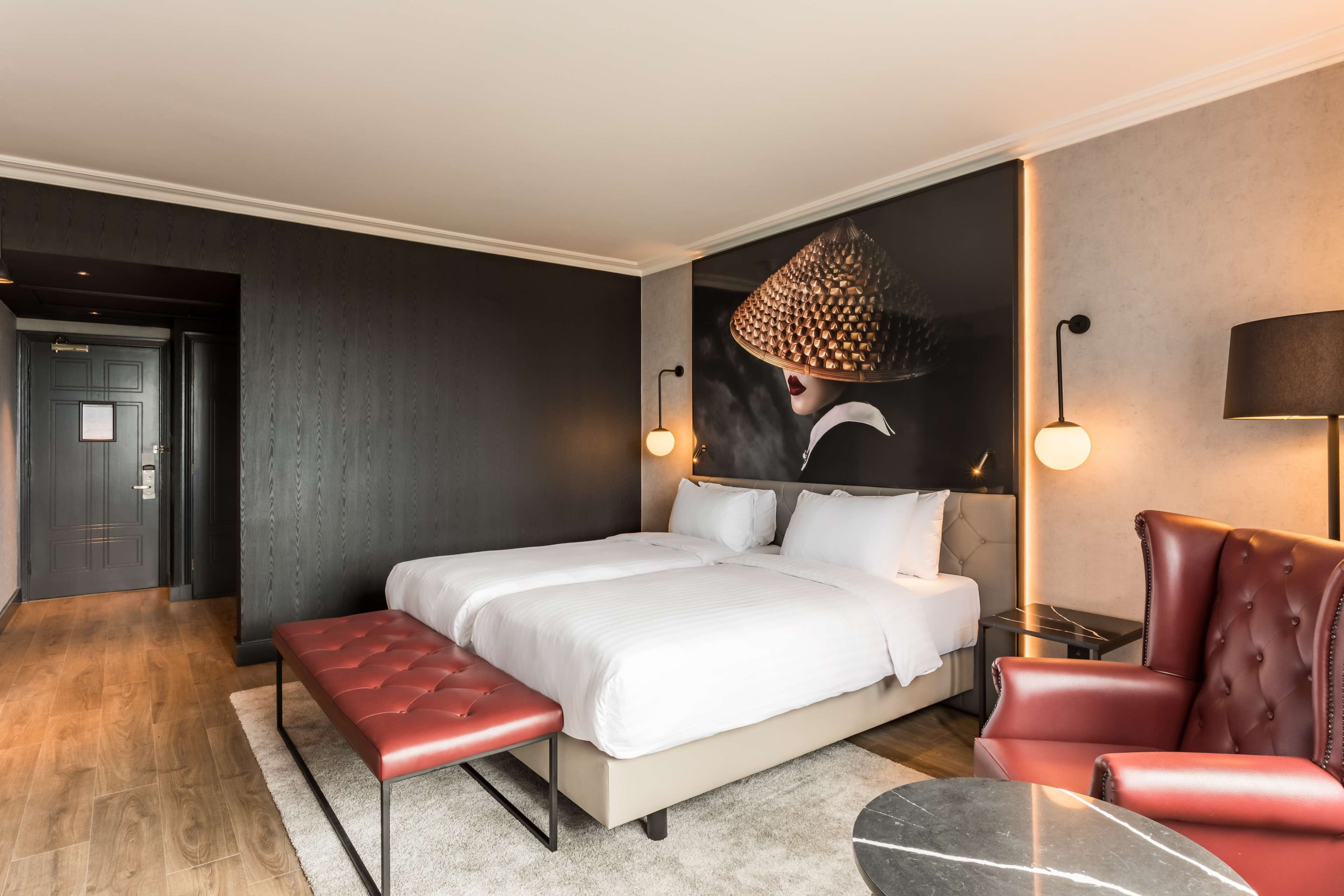 A modern hotel room with a large bed, a red leather armchair, a small round table, and a wall-mounted lamp.