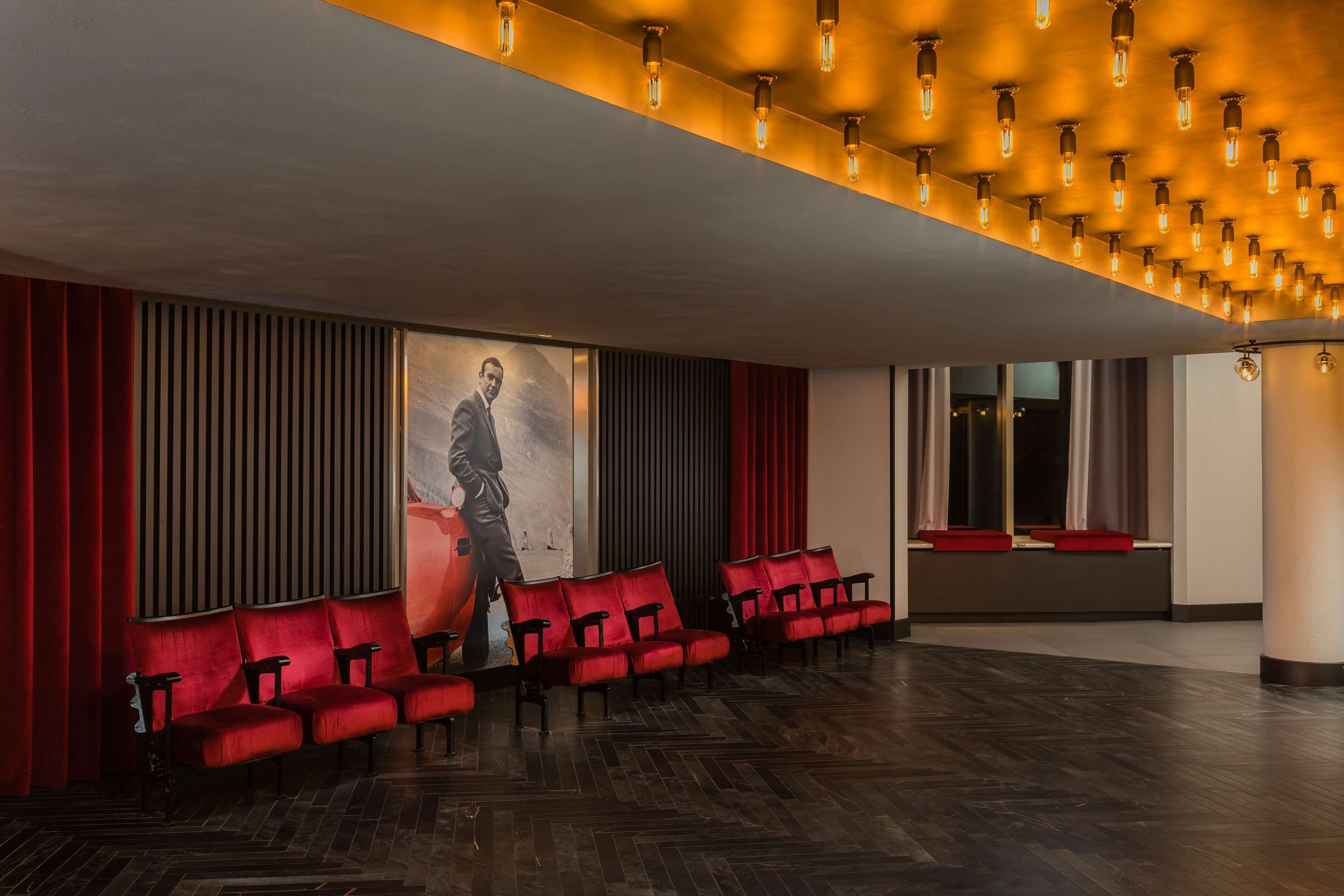 Un teatro con hileras de asientos rojos, un cartel grande en la pared y un techo con luminarias a la vista.