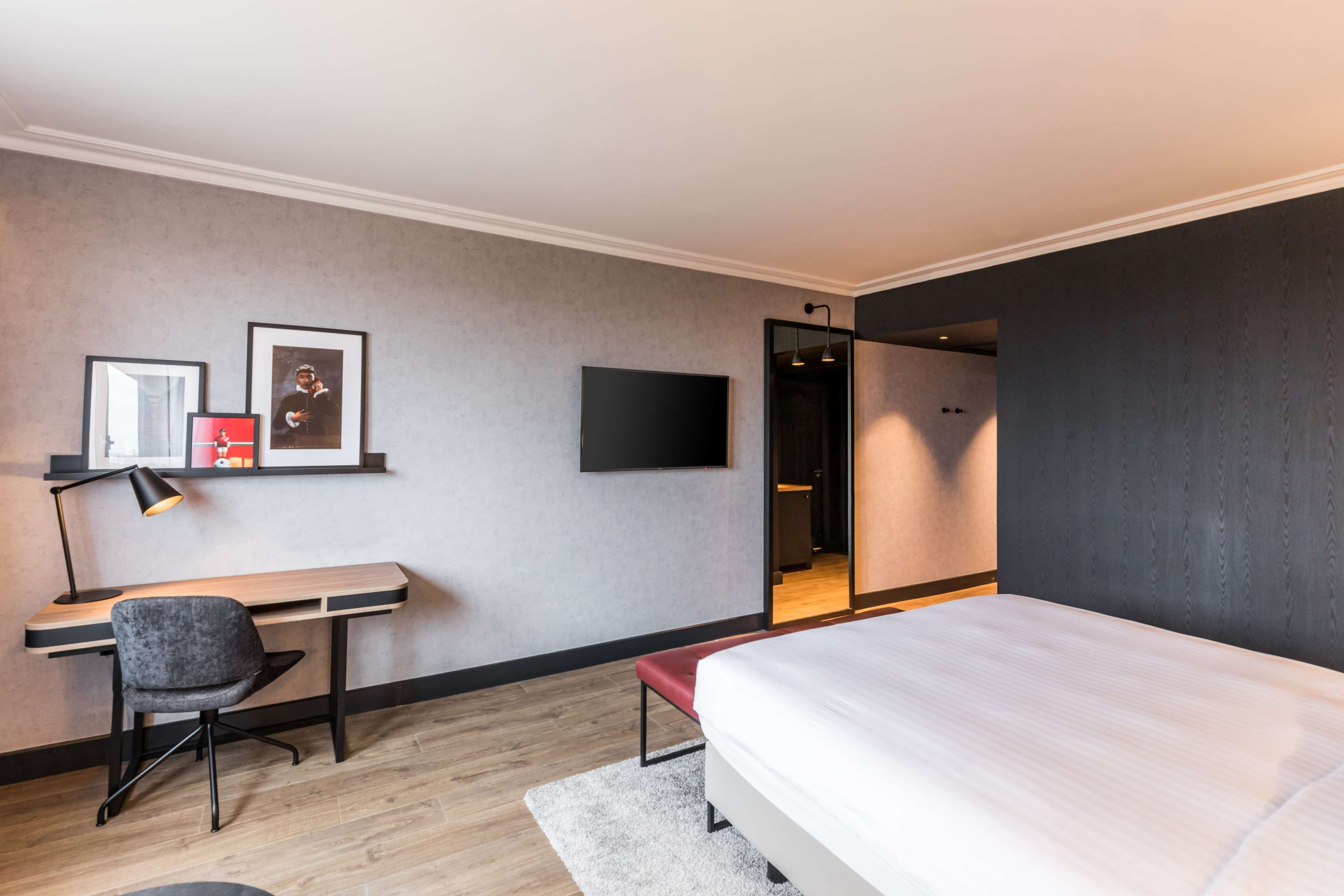 Quarto de hotel moderno com cama, escrivaninha, cadeira e televisão na parede.