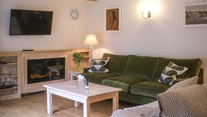 A living room with a green sofa, white armchair, wooden coffee table, fireplace, TV, wall clock, and wall-mounted lamp. - Broomhill Cottage  - sleeps 4 guests  in 2 bedrooms (Midhurst)