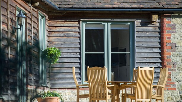 A wooden cabin with a large window, a table with chairs, and a potted plant. - Broomhill Cottage  - sleeps 4 guests  in 2 bedrooms (Midhurst)