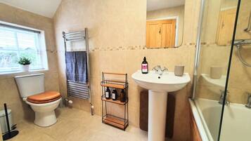 A bathroom with a toilet, towel rack, shelf with bottles, sink, and shower.