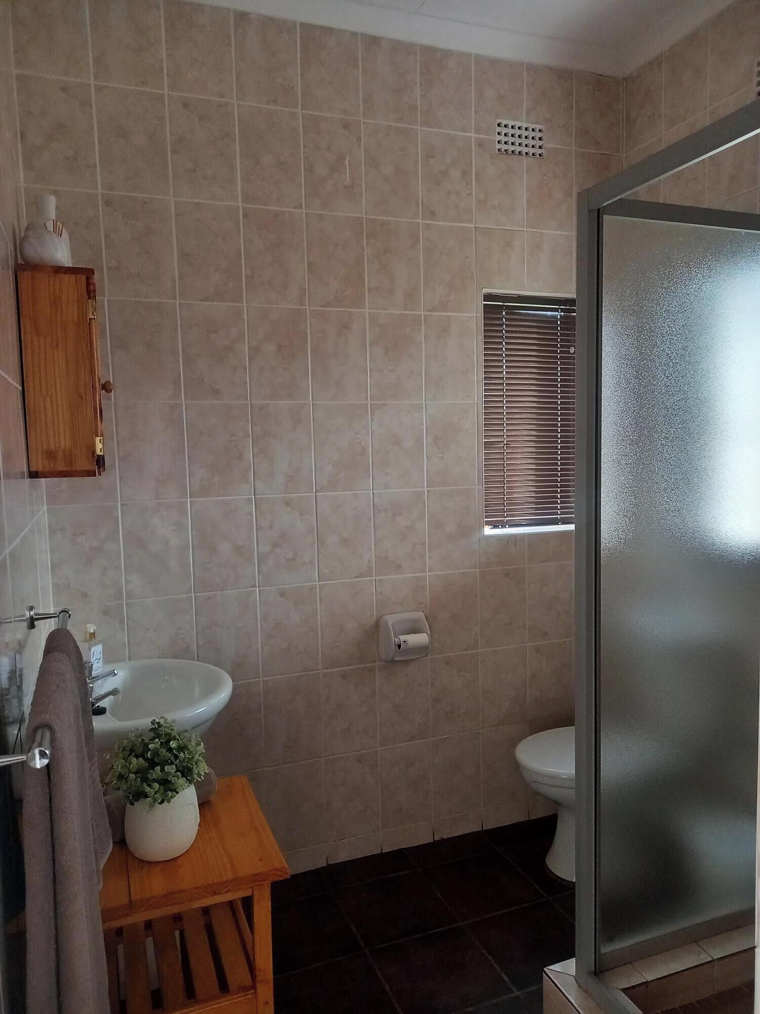 A bathroom with beige tiles, a wooden cabinet, a sink, a toilet, a window with blinds, and a shower area.