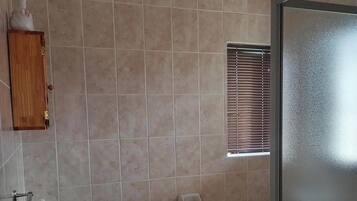 A bathroom with beige tiles, a wooden cabinet, a sink, a toilet, a window with blinds, and a shower area.