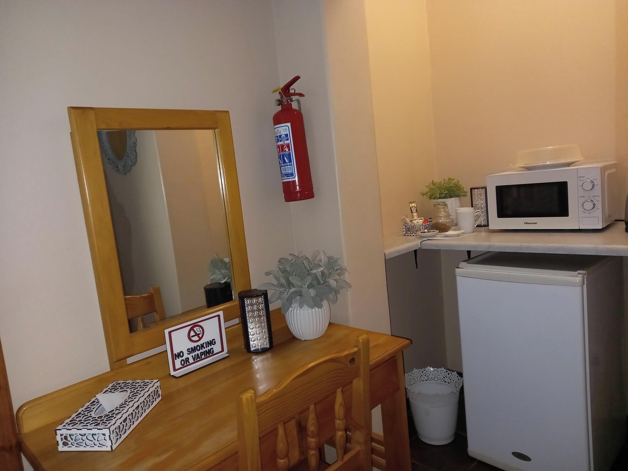 Eine kleine Küchenzeile mit einer Mikrowelle, einem Kühlschrank, einem Holztisch mit einem Schild ‚Rauchen und Dampfen verboten‘, einem Spiegel und einem Feuerlöscher.