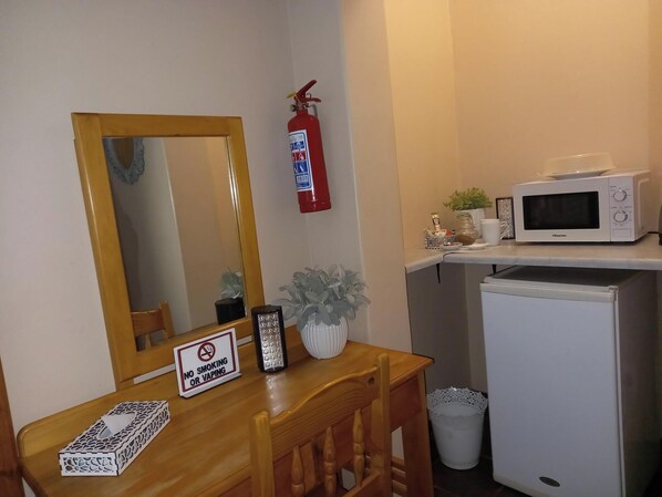 A small kitchenette with a microwave, a refrigerator, a wooden table with a 'No Smoking or Vaping' sign, a mirror, and a fire extinguisher. - Acacia Guesthouse (Kathu)