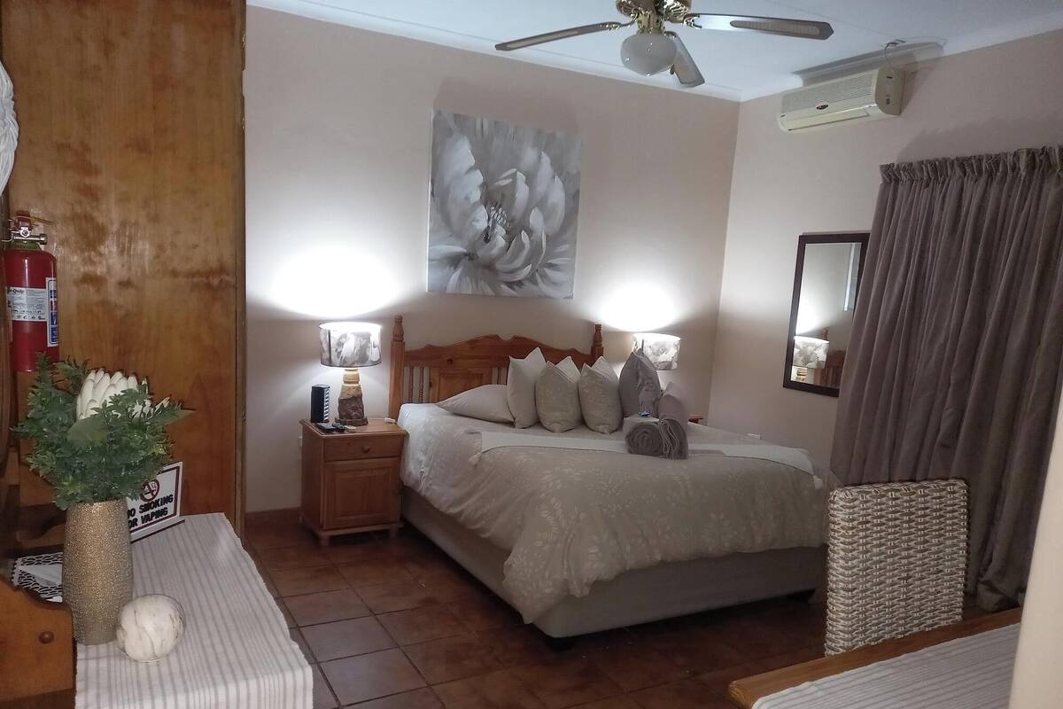 Une chambre à coucher comprenant un lit, des tables de chevet, un ventilateur de plafond, un climatiseur fixé au mur et un grand tableau représentant des motifs floraux.