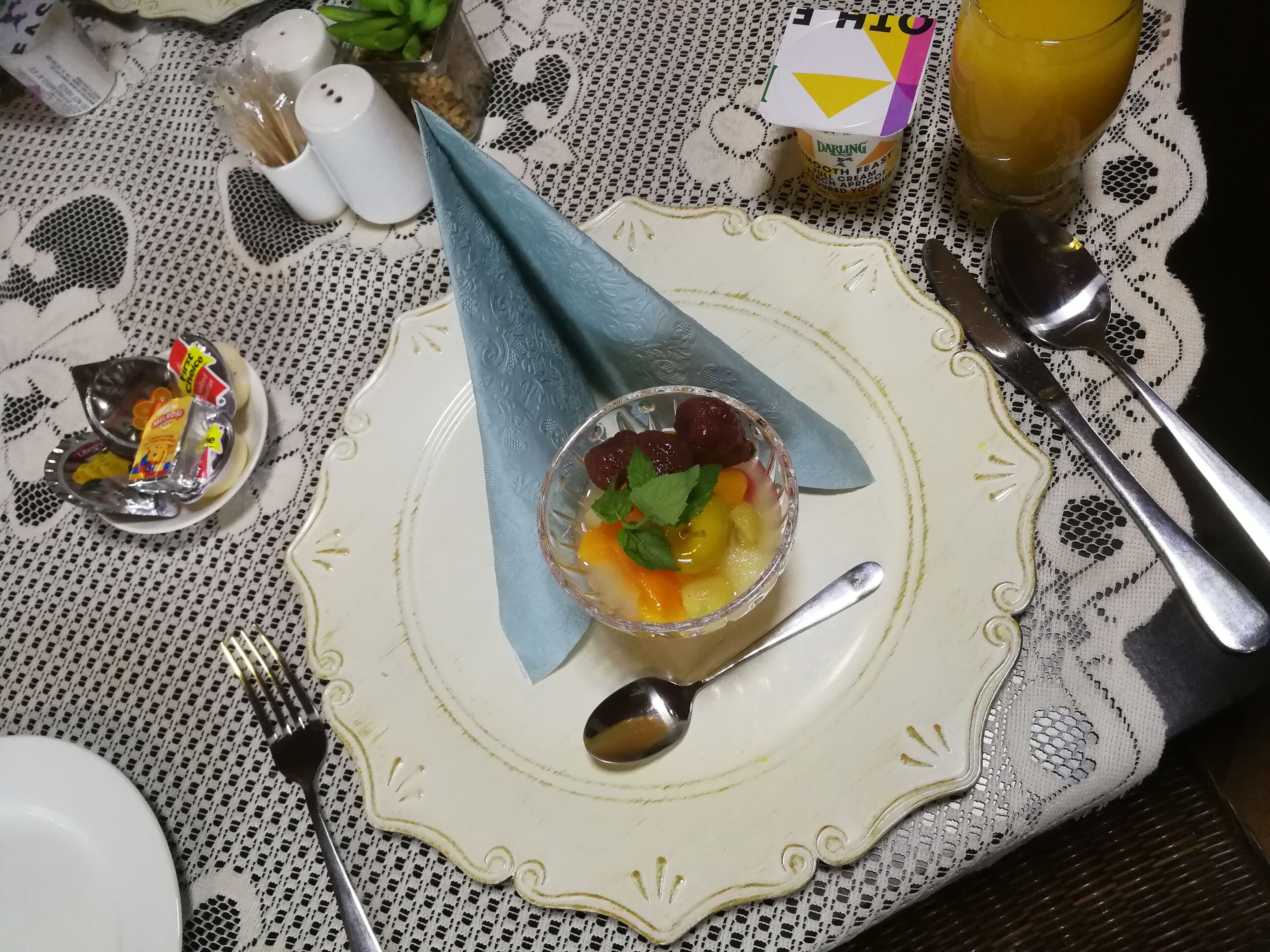 A table set with a glass bowl of fruit, a spoon, a fork, a salt and pepper shaker, a small dish with condiments, a glass of orange juice, and a napkin.