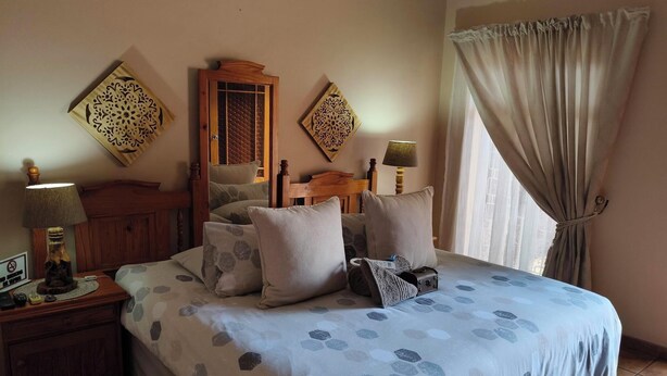 A bedroom with a bed, wooden headboard, patterned pillows, a nightstand with a lamp, and a window with curtains. - Acacia Guesthouse (Kathu)