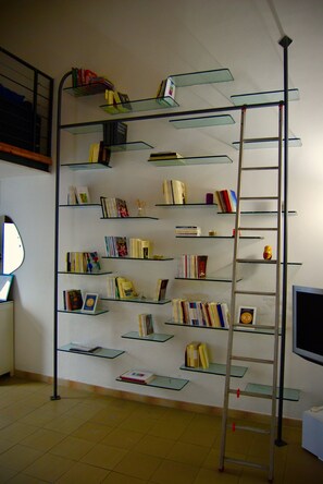 A room with a tall, narrow bookshelf filled with books, a ladder, a desk with a computer, and a television. - Open space in the center a few steps from the sea (Pescara)