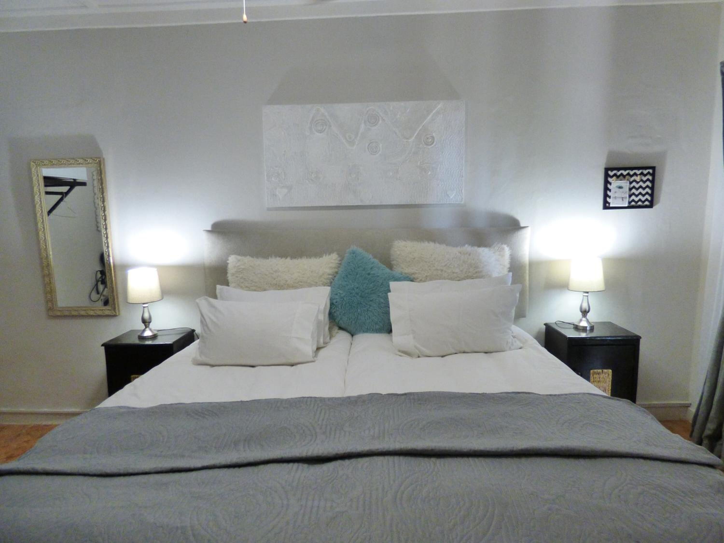 A neatly made bed with a grey blanket, white pillows, and a blue accent pillow. There are two bedside tables with lamps, a wall-mounted mirror, and a decorative piece above the bed.