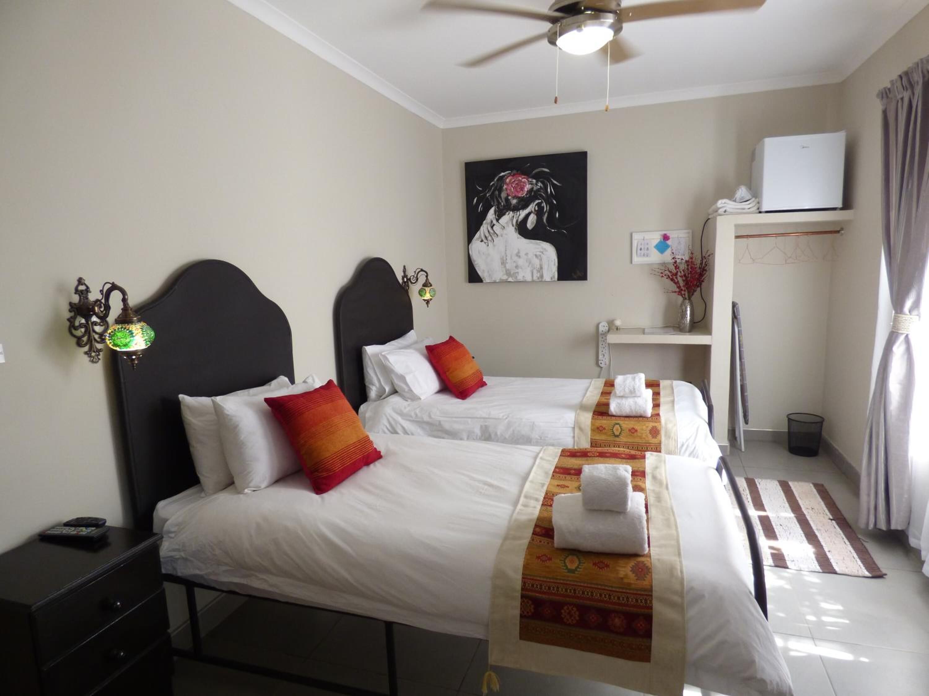 A hotel room with two beds, a ceiling fan, a painting on the wall, a desk with a computer, and a window with curtains.