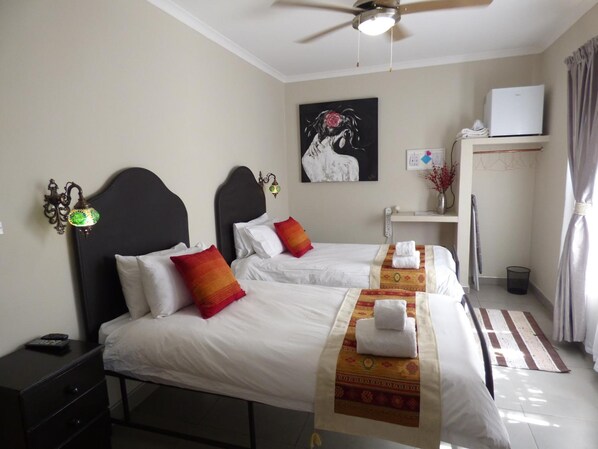 A hotel room with two beds, a ceiling fan, a painting on the wall, a desk with a computer, and a window with curtains.