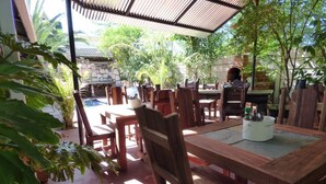 Un patio al aire libre con mesas y sillas de madera, rodeado de abundante vegetación y un muro de piedra.