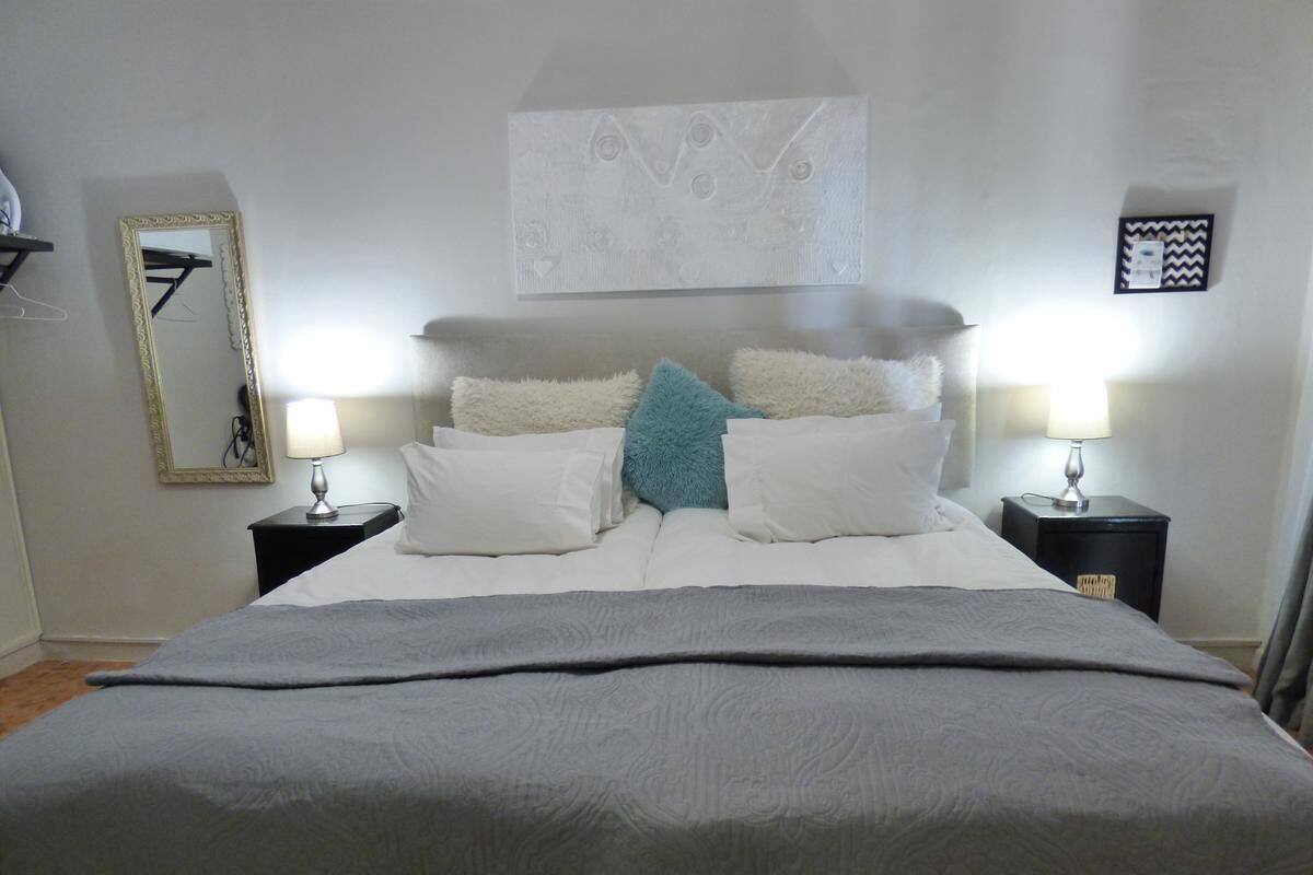 A neatly made bed with a grey blanket, two bedside tables with lamps, a mirror on the left, and a framed artwork above the bed.