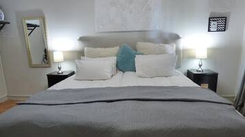 Una cama bien hecha con un edredón gris, dos mesitas de noche con lámparas, un espejo a la izquierda y una obra de arte enmarcada sobre la cama.